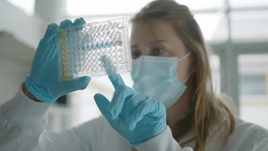 A woman in a lab coat face mask and blue gloves holds a piece of plastic and points with her finger as she holds it up in a white room with sunlight