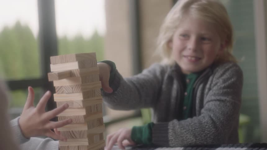 A young boy uses his finger to push a piece of wood in a puzzle on a table with two kids sitting with him as he smiles