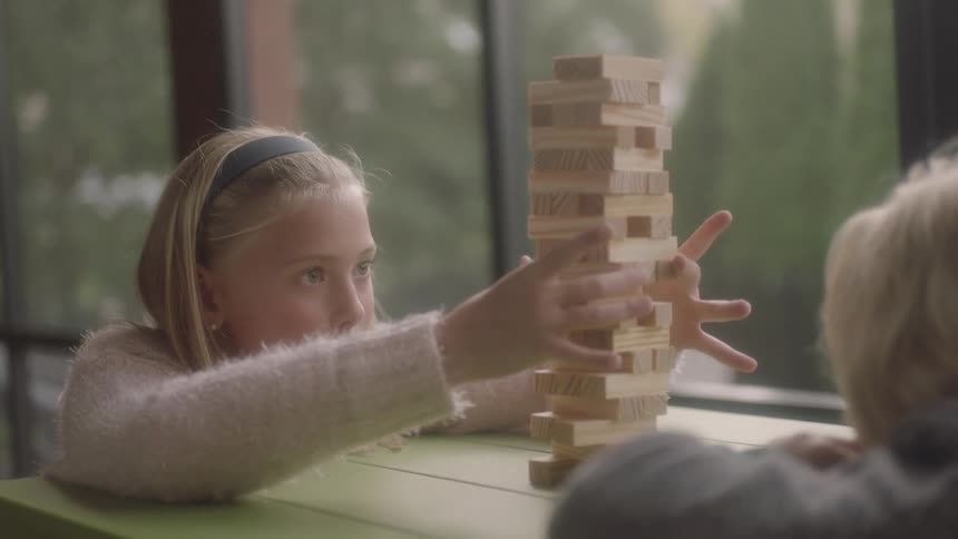 A young girl sits at a table with another child playing a game of jenga