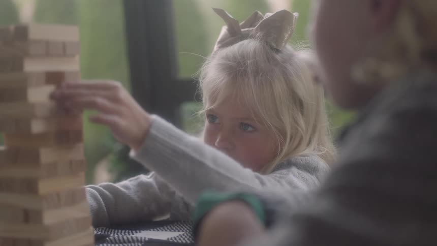 A blonde girl uses her hand to move a piece of wood on a game being played in a table with another boy inside during the day
