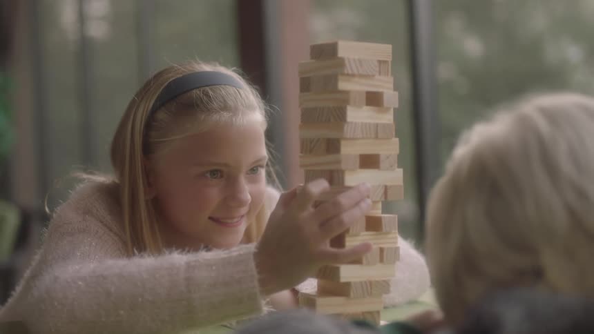 A young blonde girl sits across from another child at a table intently looking at a block of wooden pieces of a game