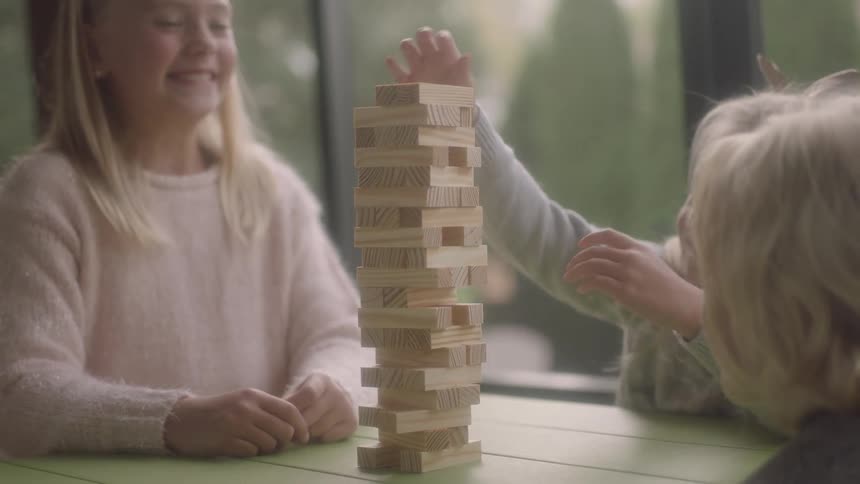 Three kids sit inside at a table playing a game with wooden pieces as they laugh