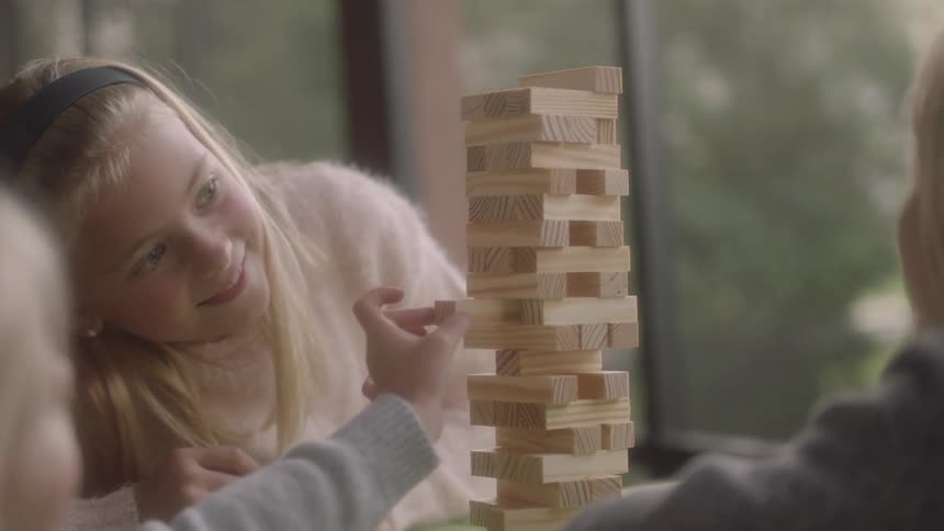 A group of children play with a jenga tower while sitting at a table near a window during the day