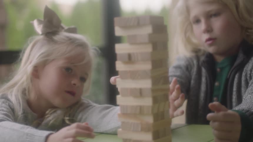 A young girl and boy play jenga at a table near a large window during the day