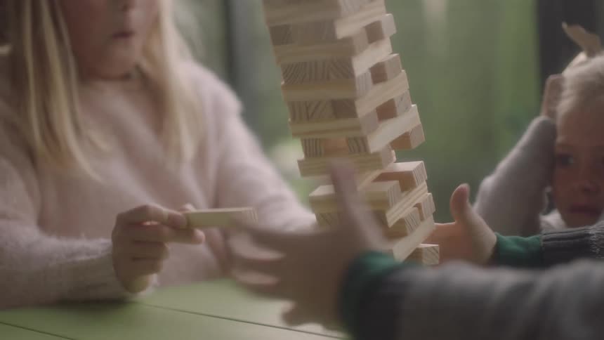 Three kids are playing a game with a tower of blocks that fall down on the table with surprised reactions