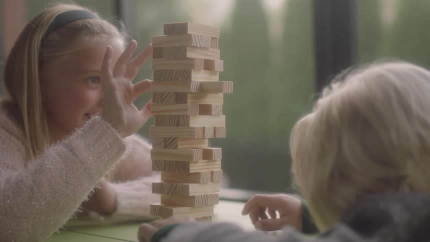 A young boy taps his fingers while waiting for his sister to remove a wooden block in a stacking game