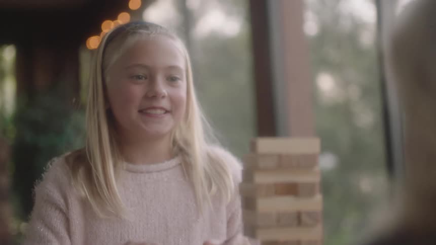 A girl smiles as she pulls a wooden block from a jenga tower before placing it at the top of the tower while playing with another girl at a table indoors during the day