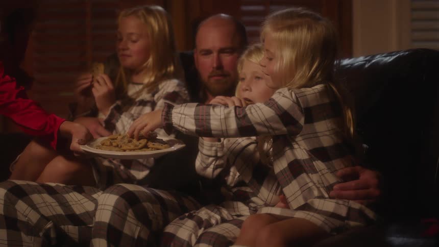 A mother holds out a plate of chocolate chip cookies for her children who sit on a sofa along with their father
