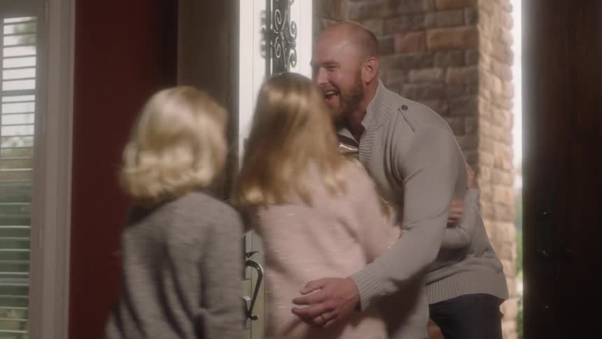 A man enters the front door of a house and is immediately greeted with hugs from three young girls