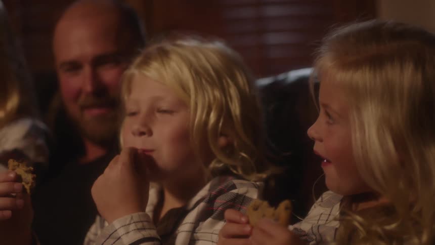 Three children sit with their dad on a couch in their pajamas eating cookies at night
