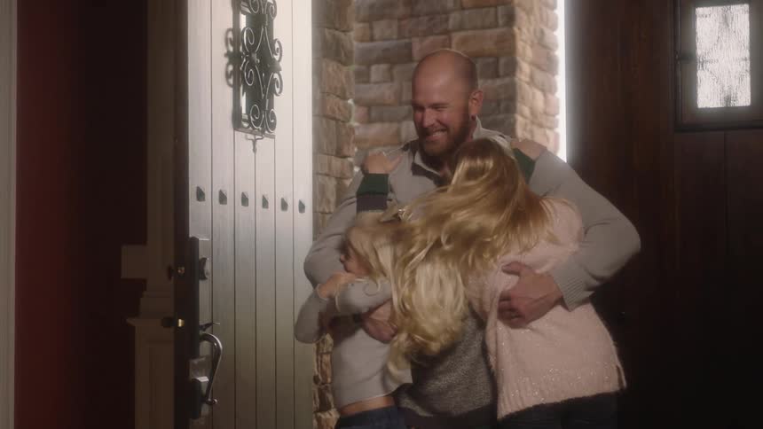 A man enters the front door of a house and is immediately greeted with hugs from his three children