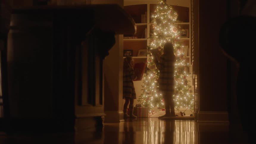 A pair of girls lift presents from beneath a christmas tree and shake them in an attempt to hear what is inside