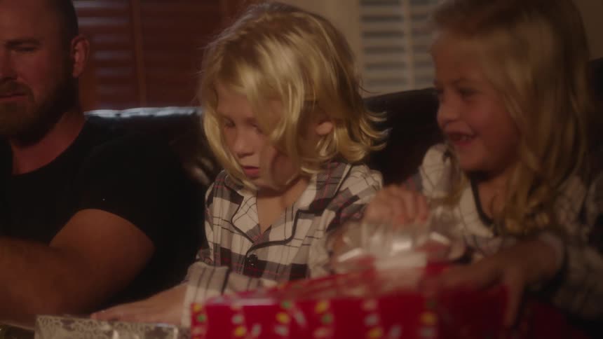 A young boy and girl open gifts while sitting on a sofa next to their father