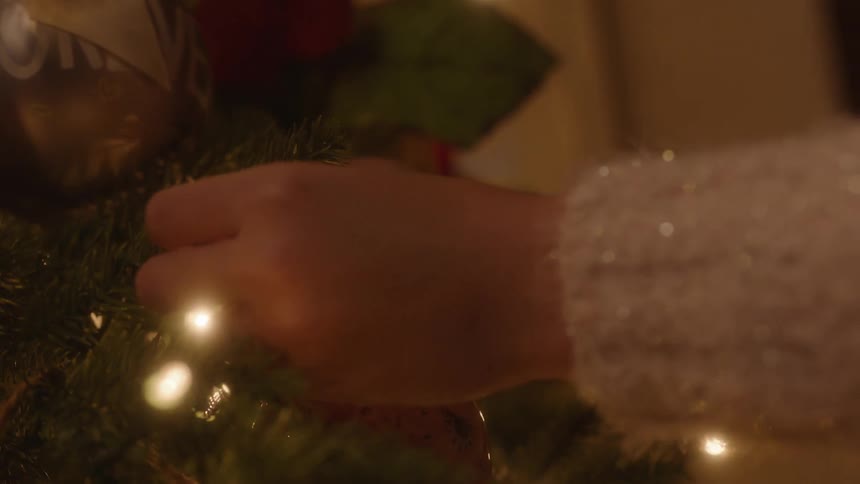 A person in a room with a sweater adding an ornament to a christmas tree with lights
