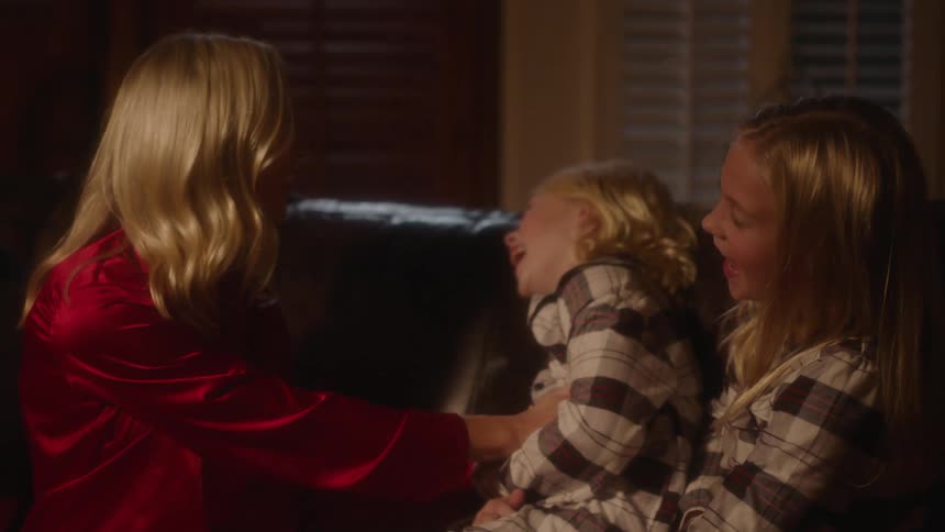 A family sits on a couch tickling each other and laughing at night in front of a christmas tree wearing pajamas