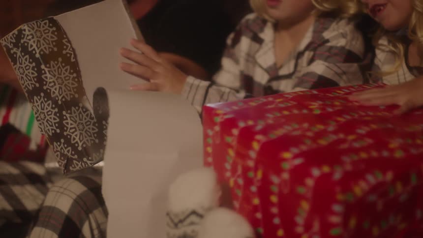 A pair of children open their gifts while sitting next to their father on a sofa
