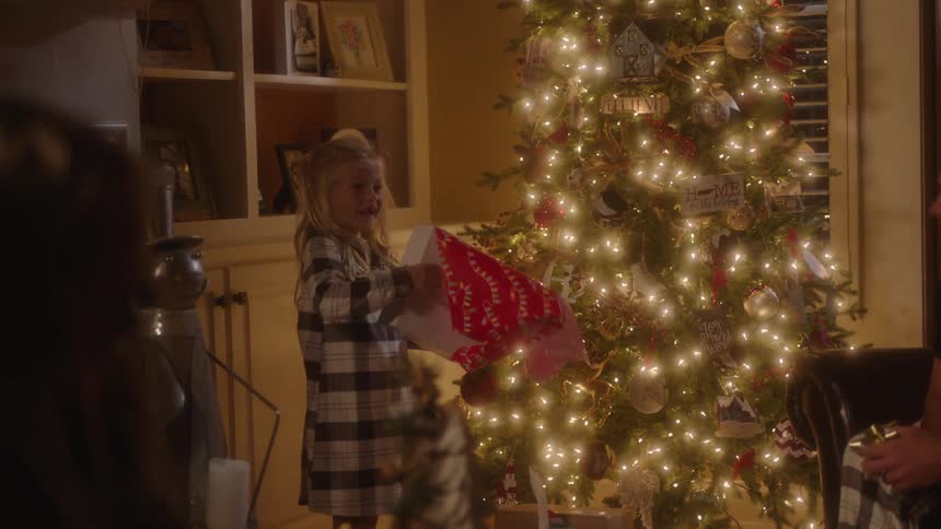 Child standing in living room in front of decorated christmas tree opening a present