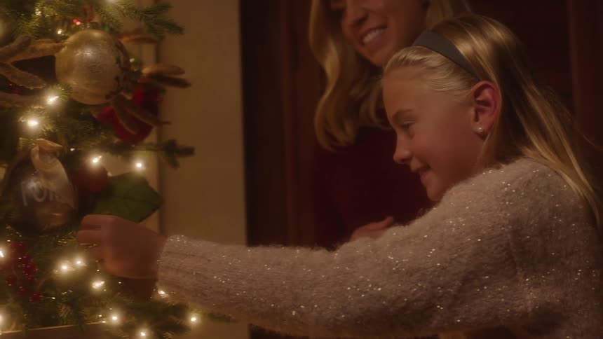 A woman hands an ornament to her daughter who places it on a christmas tree