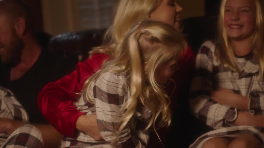 A family is sitting on a couch tickling each other and laughing while wearing pajamas at night