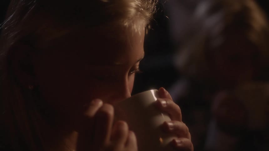 Young blonde girl drinking a hot beverage from a mug in front of a campfire at night
