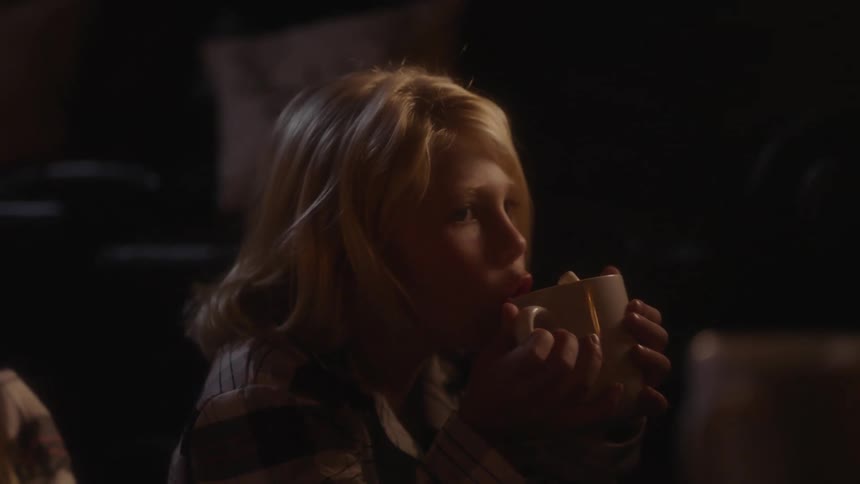 A young girl uses both hands to hold and drink from a mug while sitting by a fire indoors