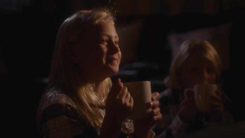Two young girls are drinking out of white mugs inside a dimly lit room