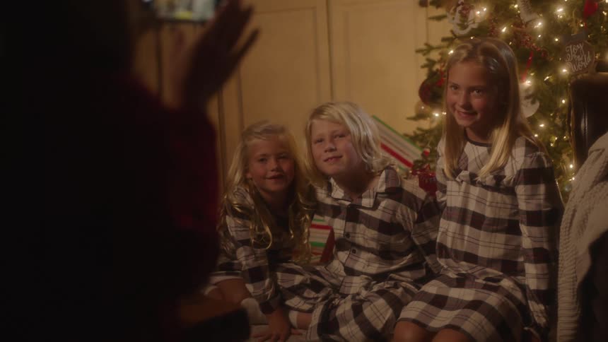 Three young siblings sit on a couch wearing matching pajamas posing for a photo in front of a christmas tree