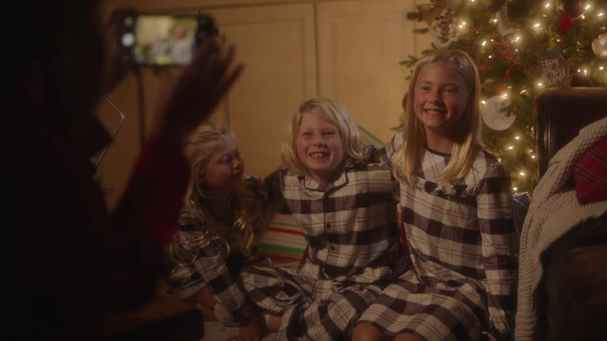 Three young siblings sit next to each other posing for a picture wearing matching pajamas in front of a christmas tree