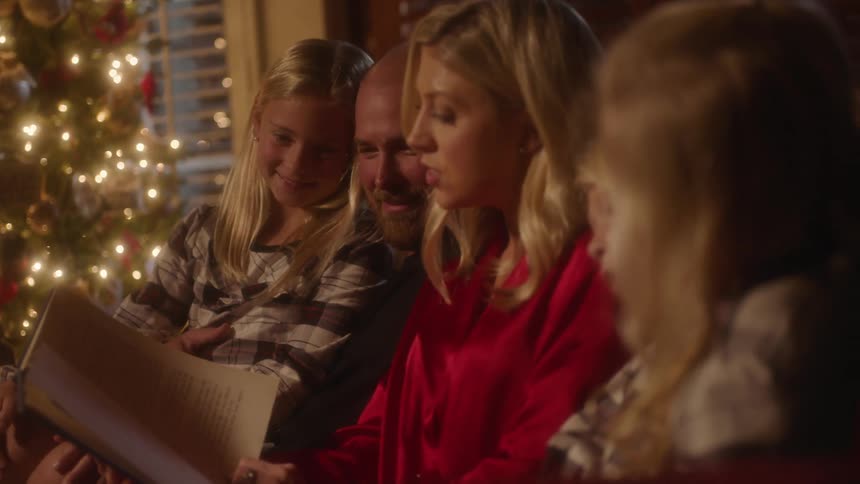 Young girls sit with their dad and mom on a couch as the mom reads from a book at night in front of a christmas tree