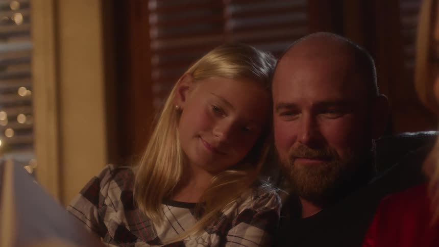 A dad and daughter sit on a couch together at night with a book in front of them