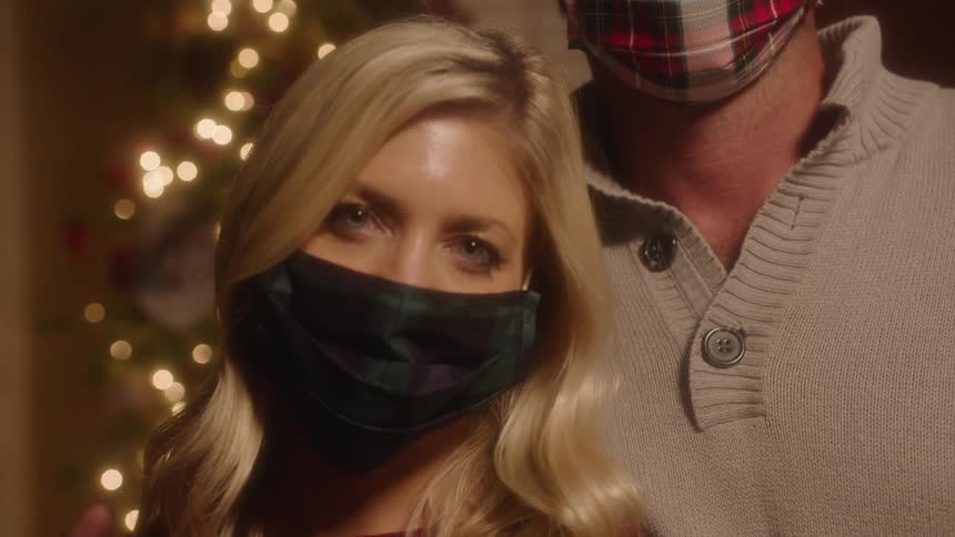 A woman in a medical mask poses in front of a christmas tree with alongside a man while gazing into the camera