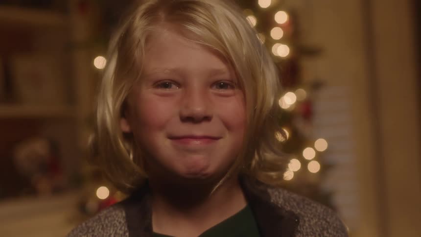 A blonde child smiles standing inside in front of a christmas tree