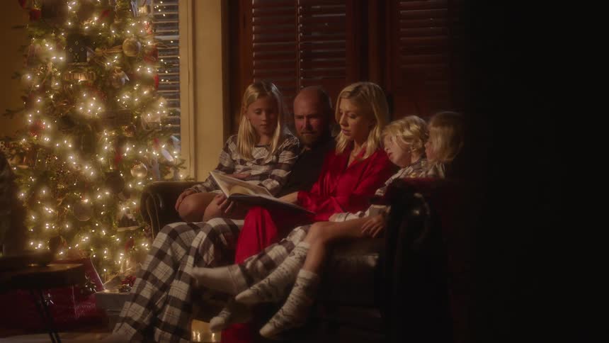 A woman reads a book aloud as she sits on a sofa with her husband and children next to a christmas tree