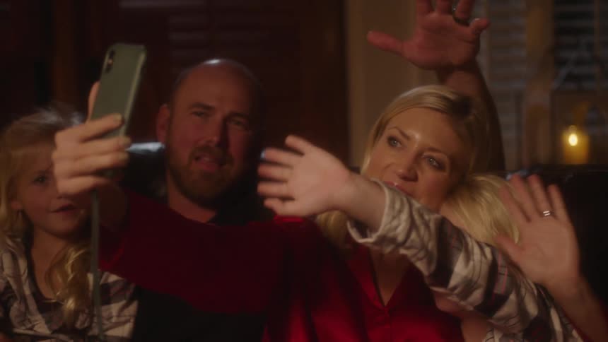 A family with three young girls a mom and a dad sit on a couch in a living room waving to a cell phone held in front of them at night