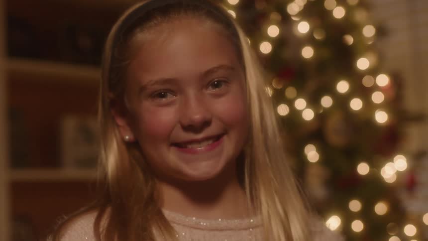 A blonde girl is smiling as she stand in front of a christmas tree indoors at night