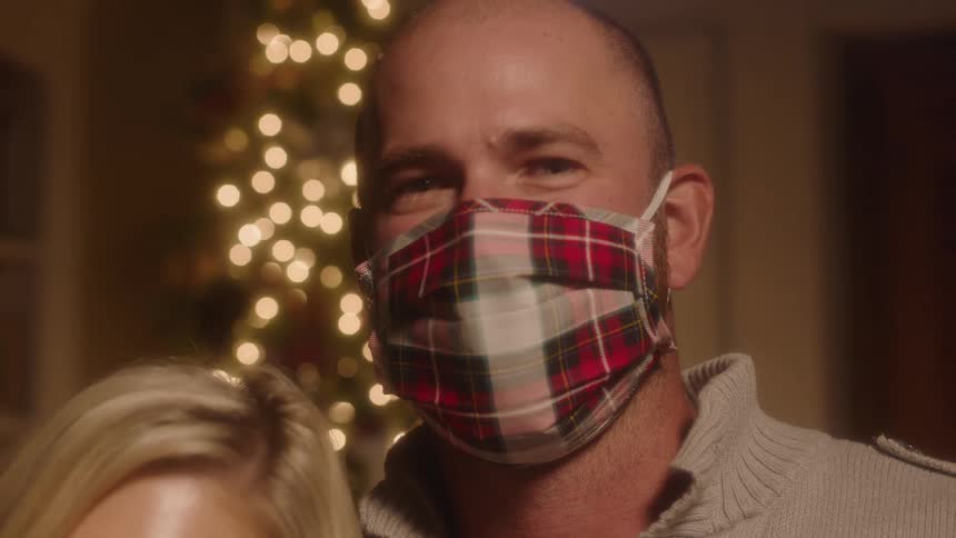 A masked smiling man stands in front of a christmas tree alongside his wife