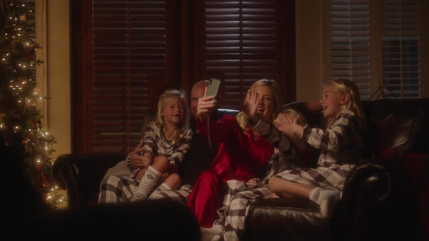 A family of mom dad and three girls sit on a couch in the living room in their pajamas waving to the phone at night