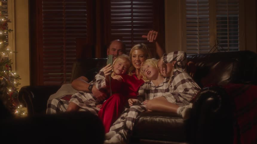 A family of give sit on their couch next to the christmas tree and wave to someone on a video call on a cell phone