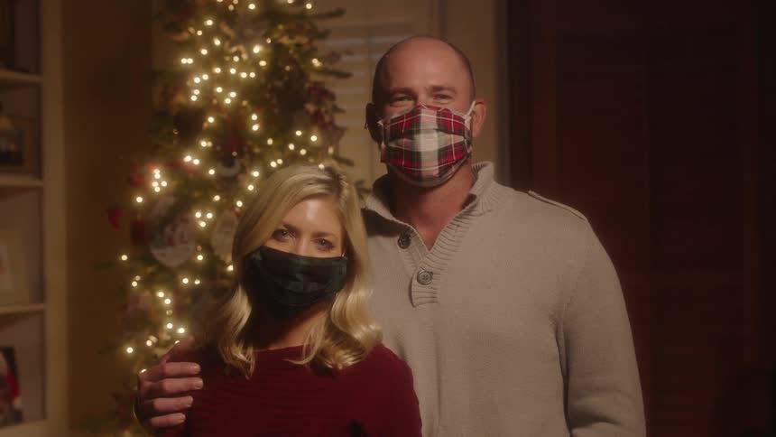A man and woman wearing masks stand side by side in front of a christmas tree while smiling