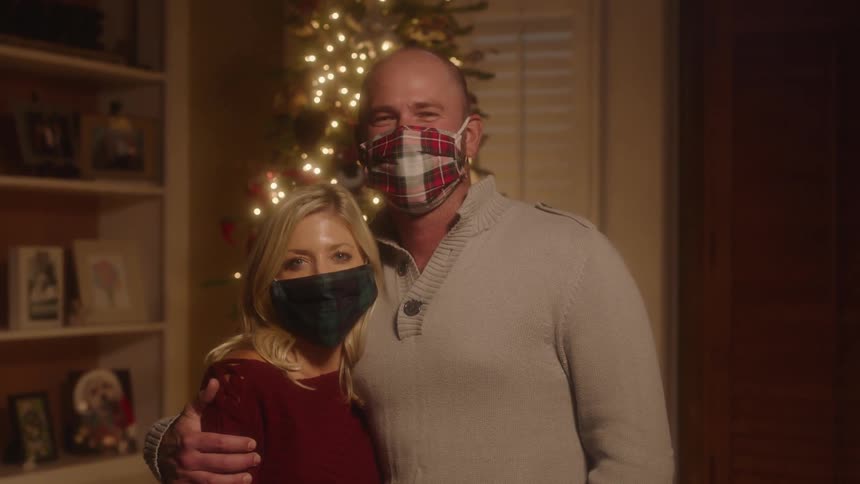 Man and woman wearing masks stand in front of christmas tree in the living room at home