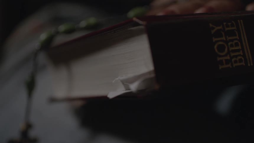 A woman's fingers can be seen resting on a bible and holding a rosary