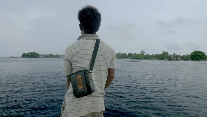 A man with a bag standing with the ocean land sky and clouds in the background