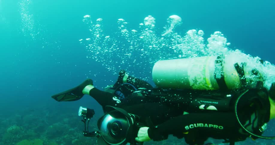 A scuba diver is floating underwater holding a large camera while bubbles rise toward the surface