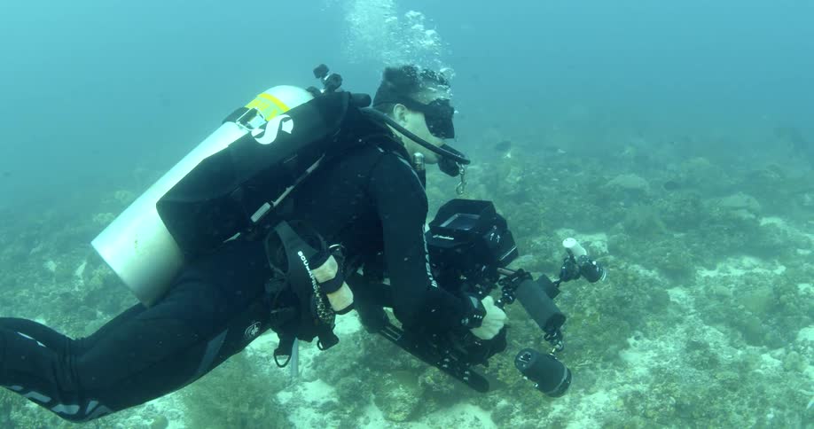 Man in snorkeling gear with oxygen tank underwater next to coral reef with fish filming with a large camera
