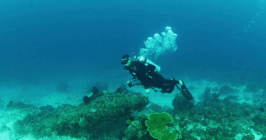 Man swims underwater in full scuba diving gear above a large coral reef