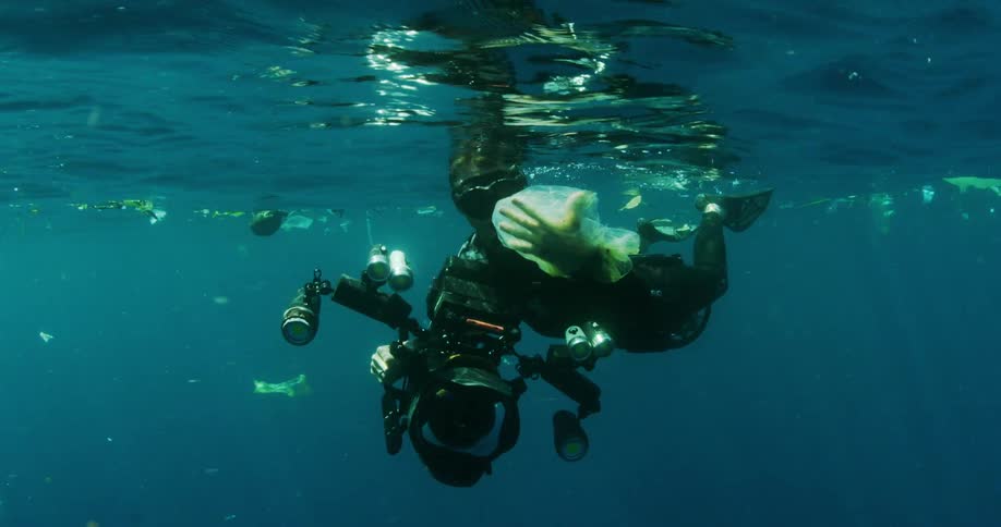 A diver with a camera is holding a plastic bag swimming through trash