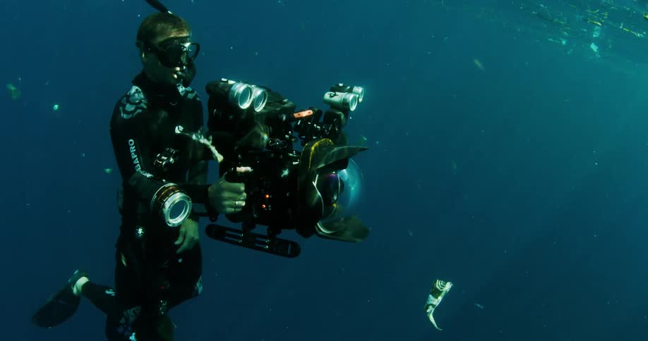 A scuba diver underwater holds a camera as he ascends to the surface of the water that is covered in trash