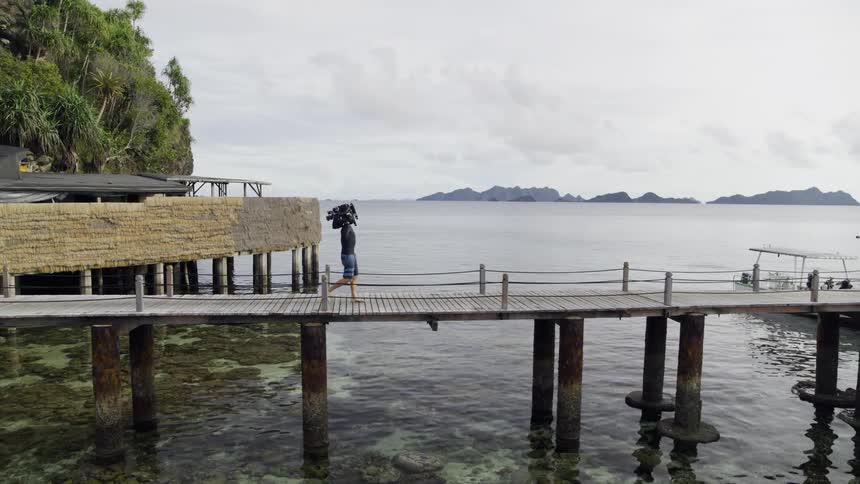 Man holding a film camera walking across a pier to a hut