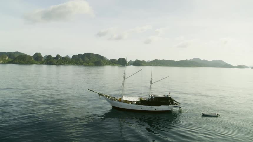 A sailboat is trailing a dinghy in the water by a coastline during the day with clear skies