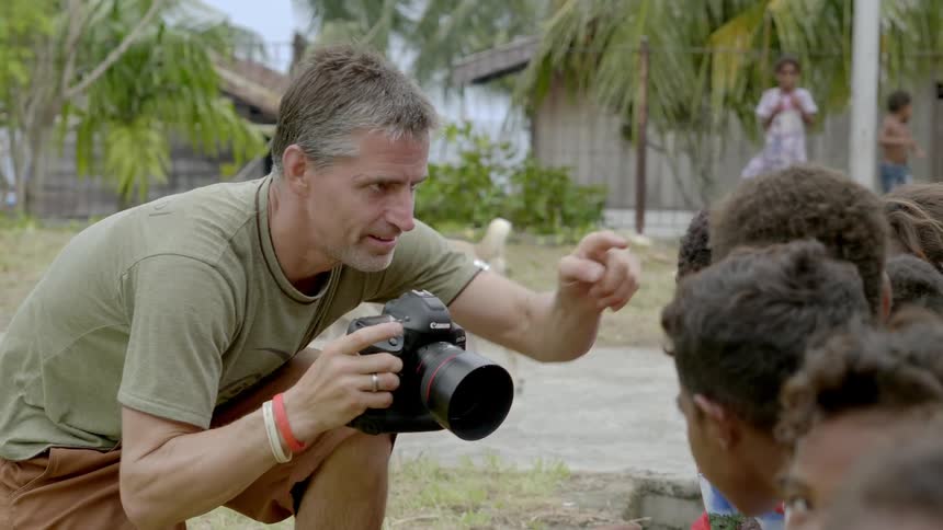 A middle aged photographer kneels to take photographs of a group of children sitting in front of him outdoors in a village